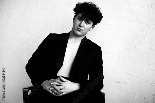 Black and white portrait of young curly man wearing black jacket, jeans, sitting, leaning on chair, interlacing fingers.