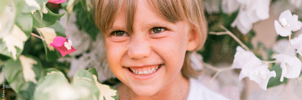portrait of face candid little happy smiling five year old blonde kid boy with green eyes in pink and white flower plants in nature. children have fun summer holidays. bright light and airy. banner portrait of face candid little happy smiling five year old blonde kid boy with green eyes in pink and white flower plants in nature. children have fun summer holidays. bright light and airy. banner