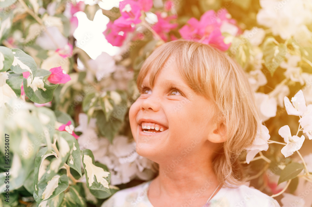 Fototapeta premium portrait of face candid little happy smiling five year old blonde kid boy with green eyes in pink and white flower plants in nature. children have fun the summer holidays. bright light and airy. flare