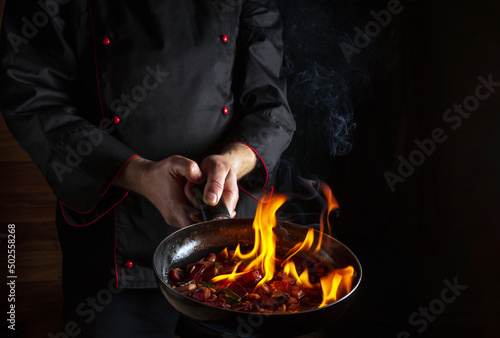 Professional chef prepares vegetable food in a frying pan with a flame of fire. The concept of restaurant and hotel service. Flamber or cooking over natural fire