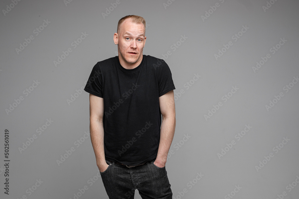 Fototapeta premium attractive man with short fair hair wearing a black t-shirt and jeans keeping hands in pockets and surprising of something