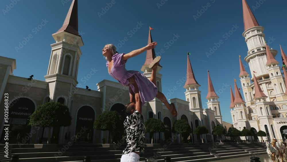Strong caucasian man holding on outstretched arms young delicate woman in pink dress while standing among popular turkish park Land of Legends. People, vacation and fun concept.