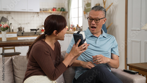 cheerful asian adult daughter surprising dad with Father’s Day gift at home. she covers his eyes and takes out the present from behind while they sit on sofa together