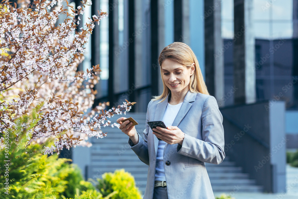 Beautiful blonde woman near a tree with flowers holds a phone and a bank credit card, smiles and makes purchases in the online store