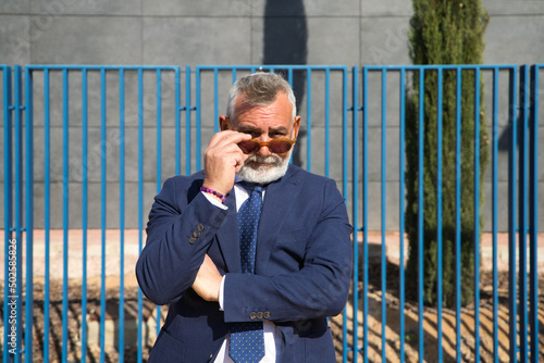 Mature, grey-haired, bearded man with sunglasses, jacket and tie makes different expressions for the photo, looks over his sunglasses and touches his lips. Concept various expressions.