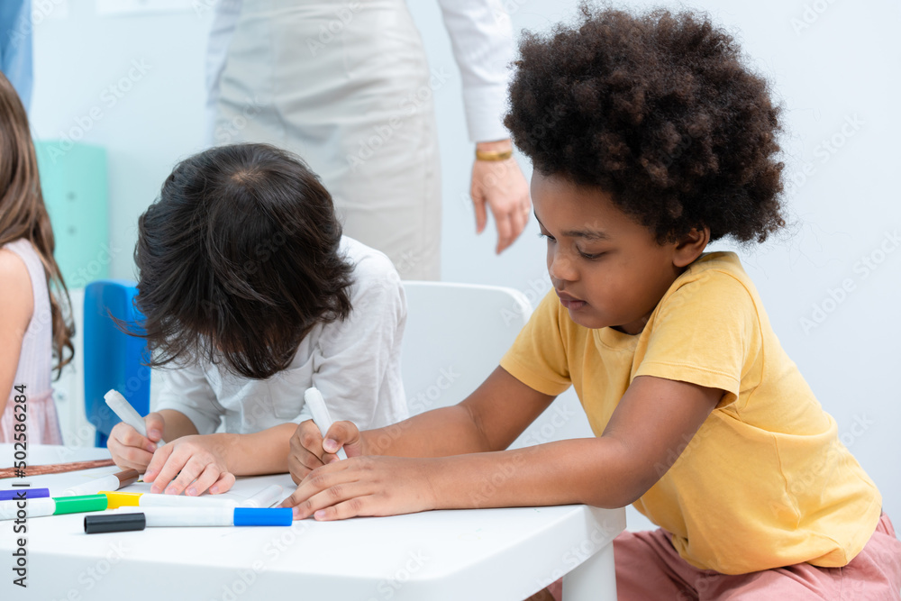 Selective focus of a little African American boy with Afro hair, sitting at a corner of table, drawing intentionally together with his friends, with a blurred teacher in the back at a kindergarten. Selective focus of a little African American boy with Afro hair, sitting at a corner of table, drawing intentionally together with his friends, with a blurred teacher in the back at a kindergarten.