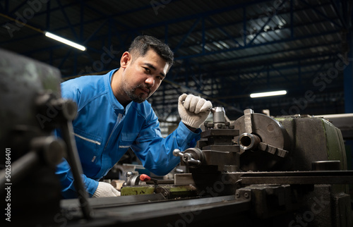 Selective focus of a professional Asian male lathe worker in a blue uniform and gloves, standing holding the tool post handle of a lathe machine while looking at the camera in a metal industry factory