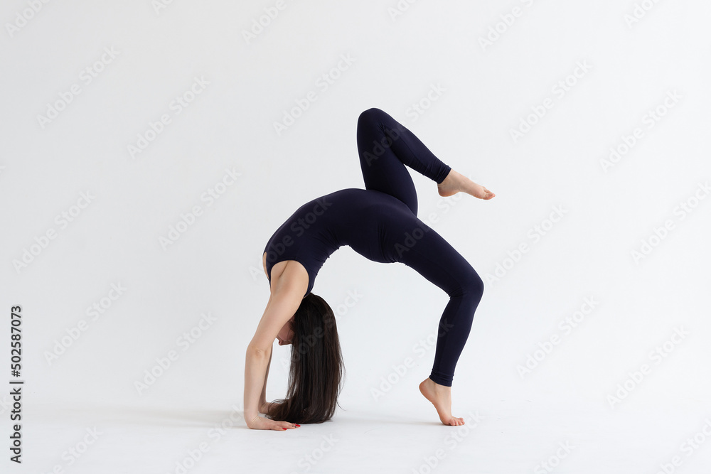 Fototapeta premium a beautiful young girl with dark hair stands in the pose of Eka Pada Urdhva Dhanurasana (variation) on a white background. Yoga class
