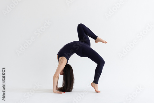 a beautiful young girl with dark hair stands in the pose of Eka Pada Urdhva Dhanurasana (variation) on a white background. Yoga class
