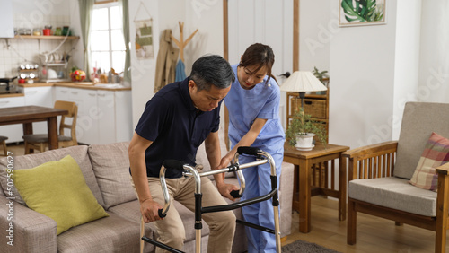 asia female nursing aide helping injured retired male to use a walker while he is slowly standing up from the couch at home. in-home health care for elderly senior concept