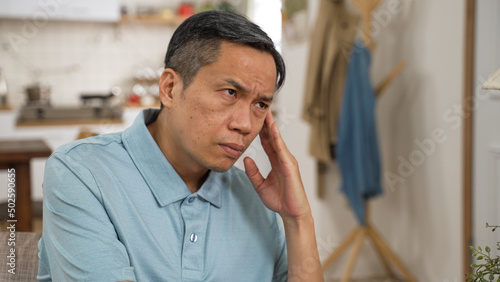 worried Japanese mature man having headache while considering hard decision alone at home. he stares into distance and shakes head feeling stressed out