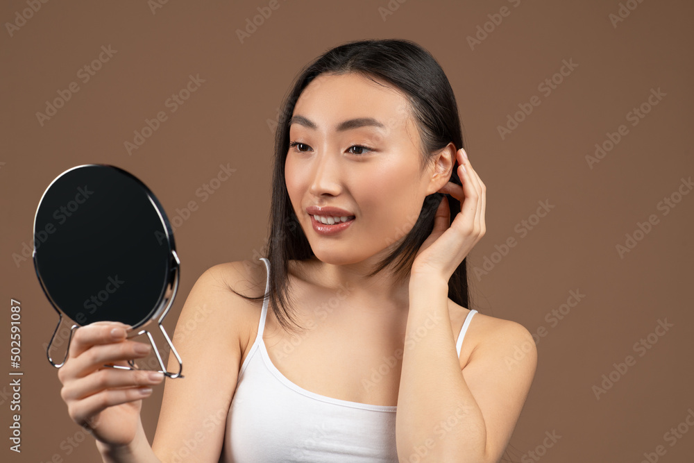 Skincare concept. Beautiful asian woman looking at mirror and touching her silky hair, posing on brown background Skincare concept. Beautiful asian woman looking at mirror and touching her silky hair, posing on brown background