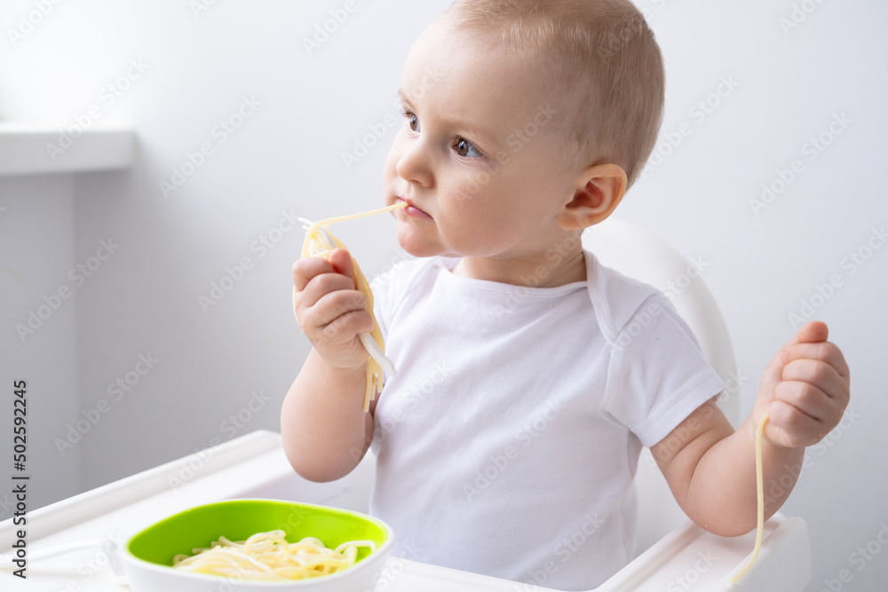 cute baby girl eating spaghetti pasta sitting in baby chair on white kitchen cute baby girl eating spaghetti pasta sitting in baby chair on white kitchen