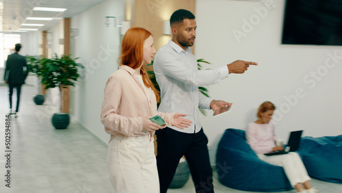 A black man is walking with a beautiful ginger woman in the office, they sit down at a seating area within the office and behind them three people are doing work at coffee tables