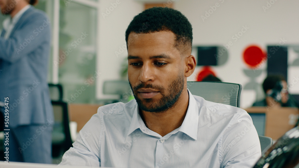Obraz premium A handsome black man with a nice haircut and a beard is sitting at the office at his desk and doing work, looking very serious