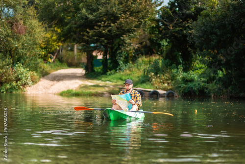 Watersport at national park. Bearded caucasian man in shirt and cap sitting in kayak and looking at paper map. The concept of the World Tourism Day © _KUBE_