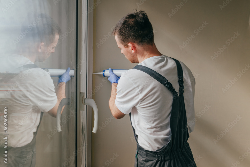 Young man wearing overalls sealing cracks between window and trim Young man wearing overalls sealing cracks between window and trim