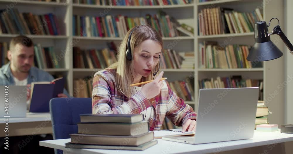 Beautiful blonde female in headset talking, writing and videochatting on laptop in library. Woman speaking, taking notes and having videochat online via webcam. Video call. Noting information. Beautiful blonde female in headset talking, writing and videochatting on laptop in library. Woman speaking, taking notes and having videochat online via webcam. Video call. Noting information.