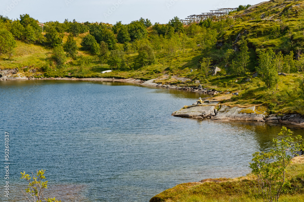 A person sits on a stone and enjoys the beauty, the picturesque coast of the mountain lake of Norway near the Lofoten Islands, the slopes of the mountains covered with green grass A person sits on a stone and enjoys the beauty, the picturesque coast of the mountain lake of Norway near the Lofoten Islands, the slopes of the mountains covered with green grass