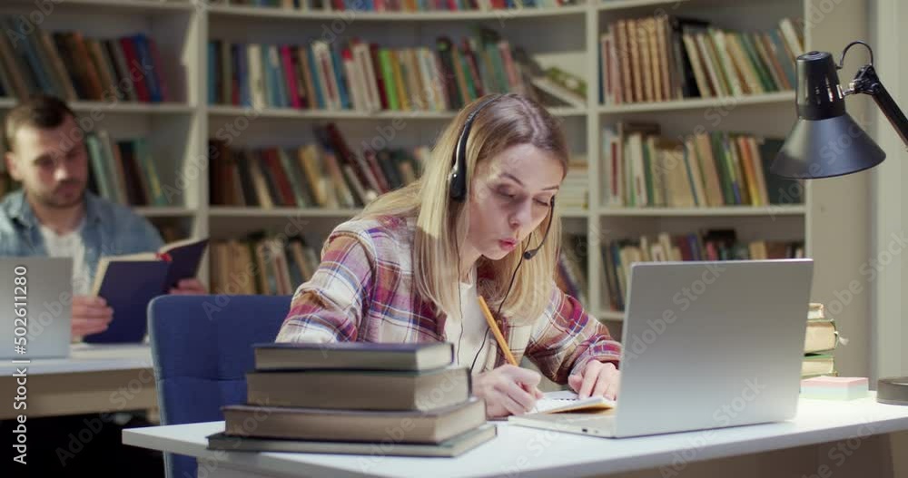 Pretty blond female in headset talking, writing and videochatting on laptop in library. Attractive woman speaking, taking notes and having videochat online via webcam. Video call. Noting information. Pretty blond female in headset talking, writing and videochatting on laptop in library. Attractive woman speaking, taking notes and having videochat online via webcam. Video call. Noting information.