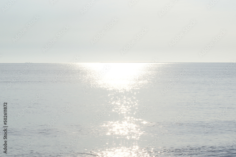 Light silvery shiny ocean background with the sun reflecting off the water in the center. Both the sky and water are in gray and white tones making this a perfect backdrop for banners and promos.