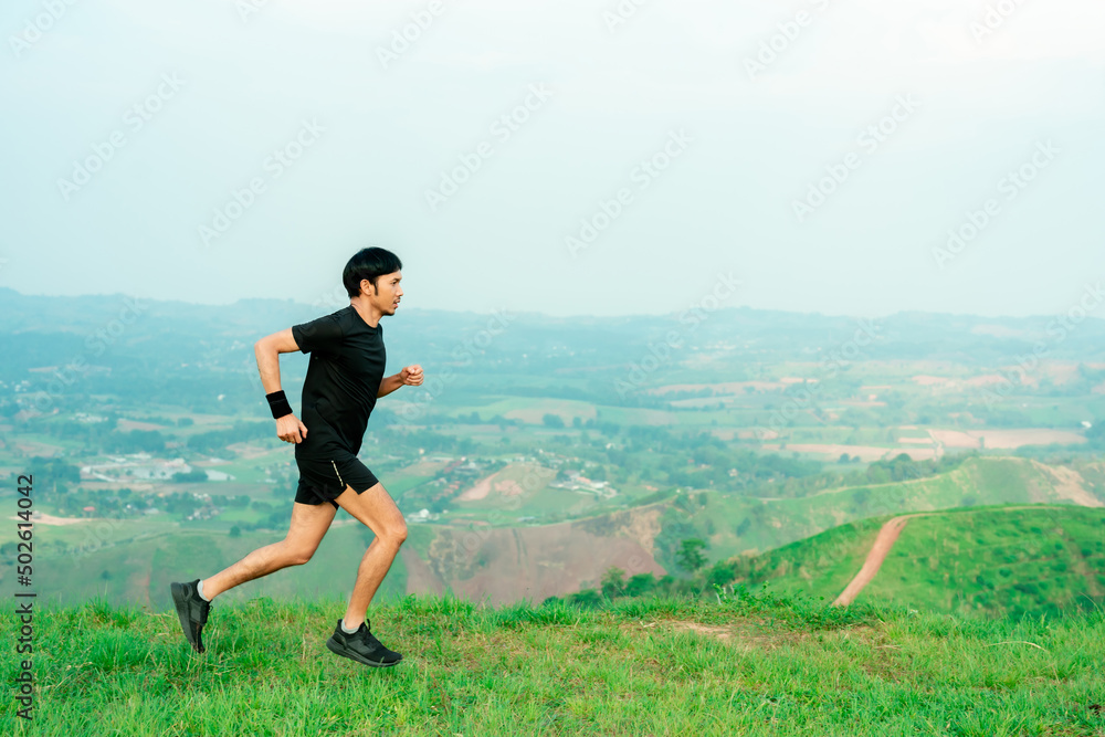 Fototapeta premium Young Asian man runner, wearing black sportswear, running on a big mountain trail, cool morning, windmills, and sky in the background.