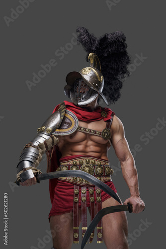 Shot of muscular ancient gladiator dressed in red cape with armor and helmet holding two swords.