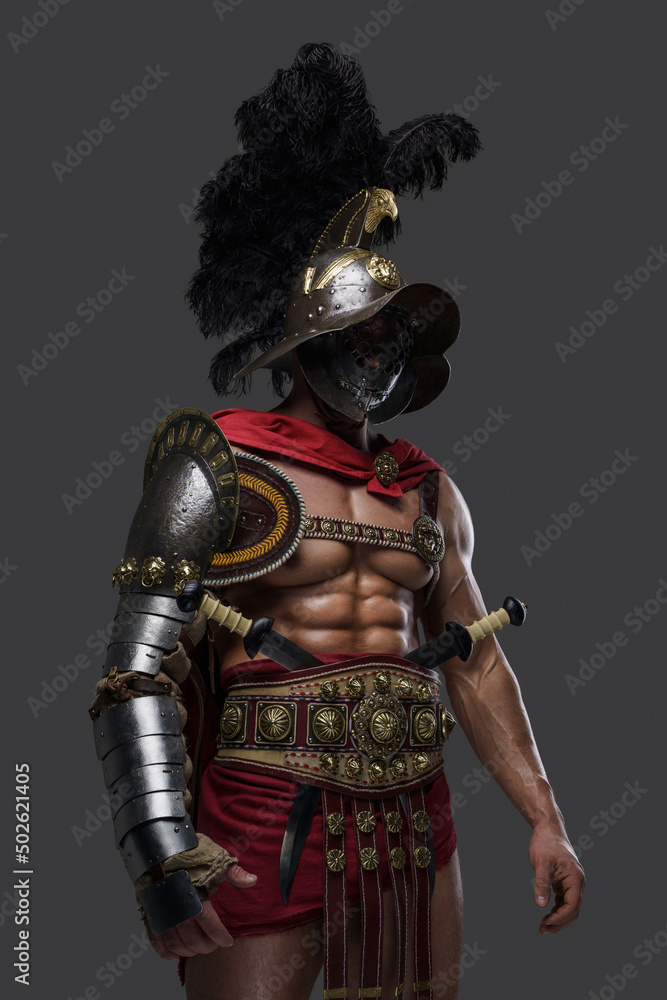Fototapeta premium Shot of violent roman gladiator dressed in light armor and red cape against gray background.