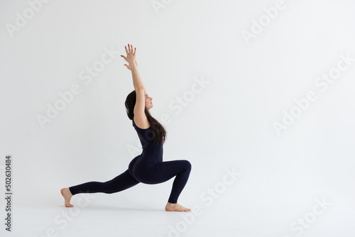 a beautiful young girl with dark hair stands in the pose of Virabhadrasana 1 on a white background. Yoga class