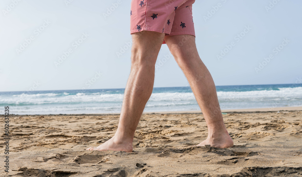 Photo of male legs wearing a swim suit while walking barefoot at the seaside of the beach in summer.
