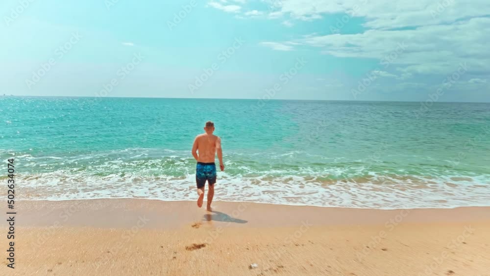 Slow-motion back view tracking shot of young man running along a sandy beach into the sea with clear blue water. Summer vacation on the sea concept. Slow-motion back view tracking shot of young man running along a sandy beach into the sea with clear blue water. Summer vacation on the sea concept.