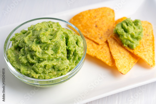Close up view of Mexican guacamole homemade vegetable avocado-based dip, spread, or salad made of mashed ripe green avocado served in glass bowl on plate with tortilla chips or nachos as appetizer