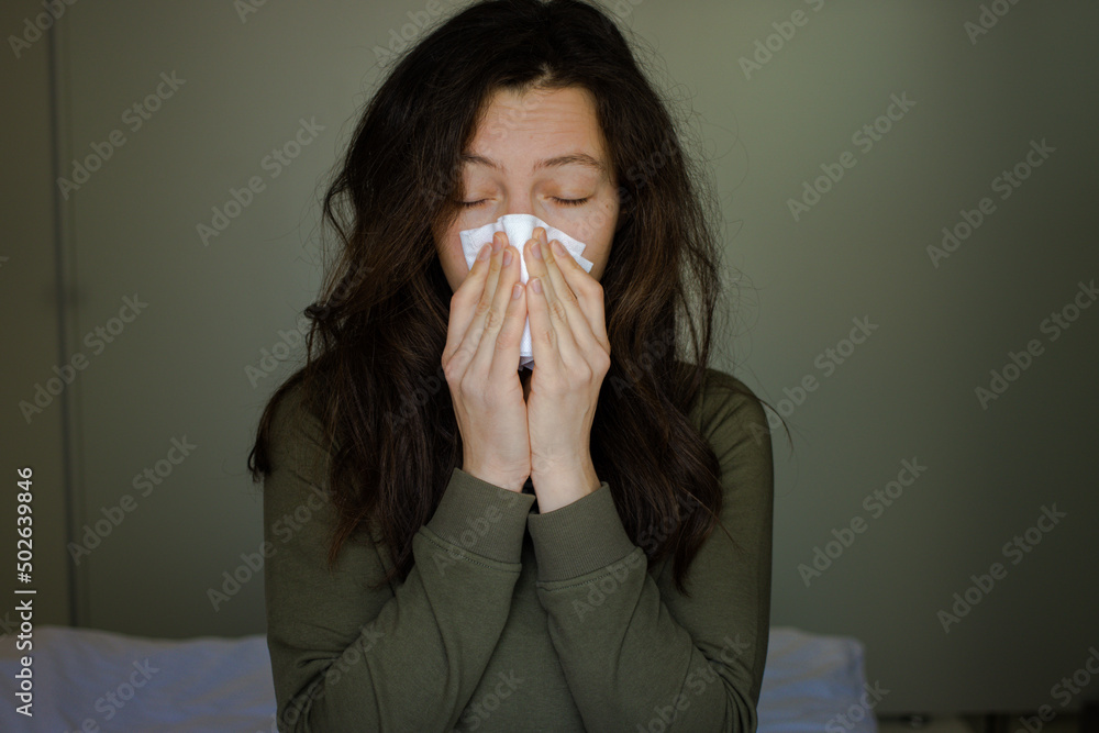 Young brunette woman sneeze into paper napkin, blowing running nose reducing breathing relief discomfort, sitting in bed at home. Seasonal pollen allergy, nasal mucosa inflammation, viruses infection Young brunette woman sneeze into paper napkin, blowing running nose reducing breathing relief discomfort, sitting in bed at home. Seasonal pollen allergy, nasal mucosa inflammation, viruses infection