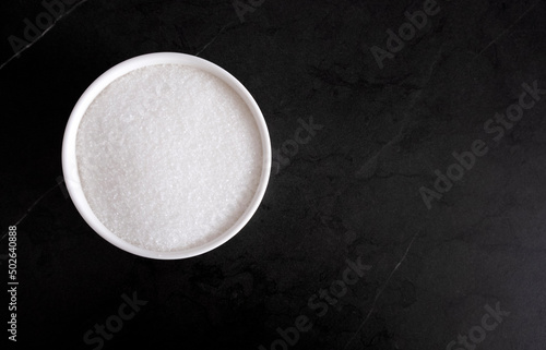 Granules of white sugar in a white bowl on a black background, free space