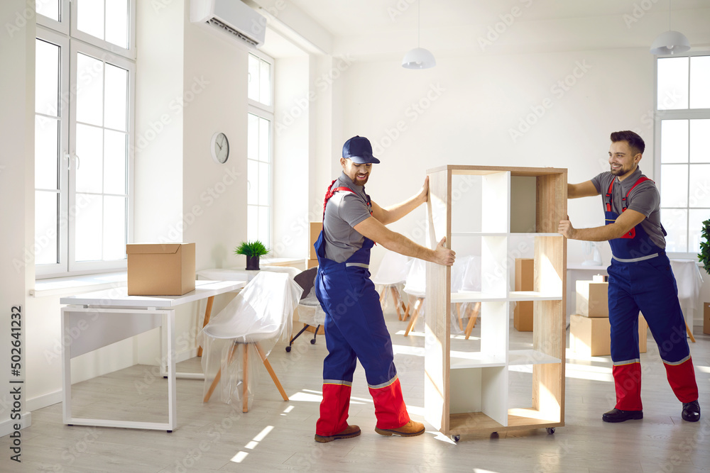 Fototapeta premium Smiling carriers help customer with furniture during relocation and moving day. Deliverymen or movers in uniform carry shelves work for client at office or new home settle. Transportation concept.
