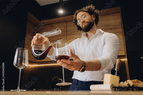 Curly man at home in a beautiful kitchen pours red wine into a glass with decanters. A sommelier pouring red wine into decanter photo