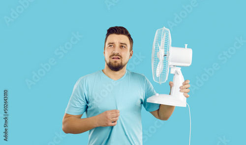 Man suffers from summer heat at home. Guy with broken air conditioner in his house using bad electric fan and sweating. Man in T shirt feeling hot and holding fan with sad face expression, studio shot photo