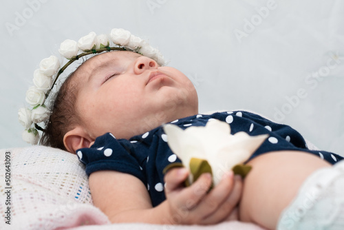 close-up detail of the face of a beautiful latin baby girl resting on a white pillow. holding in her right hand a white flower symbol of her purity and innocence. concept of innocence. photo