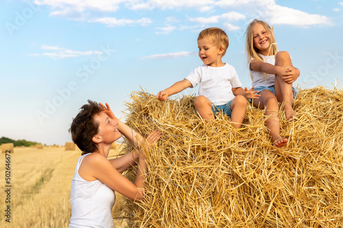 Valokuvatapetti Young adult attractive beautiful mom with little son and daughter enjoy having fun fooling around sitting near golden hay bale on wheat harvested field near farm
