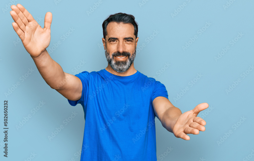 Middle aged man with beard wearing casual blue t shirt looking at the camera smiling with open arms for hug. cheerful expression embracing happiness. Middle aged man with beard wearing casual blue t shirt looking at the camera smiling with open arms for hug. cheerful expression embracing happiness.