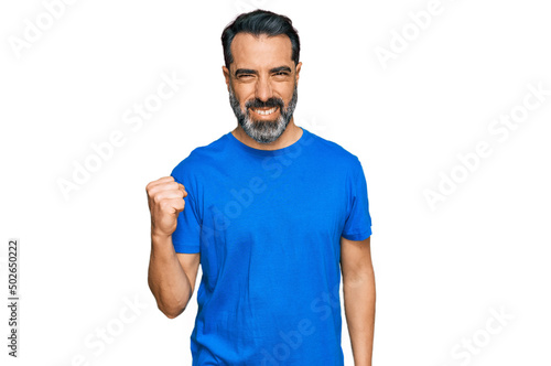 Middle aged man with beard wearing casual blue t shirt angry and mad raising fist frustrated and furious while shouting with anger. rage and aggressive concept.