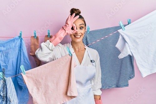 Beautiful brunette young woman washing clothes at clothesline doing ok gesture with hand smiling, eye looking through fingers with happy face.