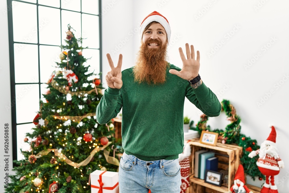 Redhead man with long beard wearing christmas hat by christmas tree showing and pointing up with fingers number seven while smiling confident and happy. Redhead man with long beard wearing christmas hat by christmas tree showing and pointing up with fingers number seven while smiling confident and happy.