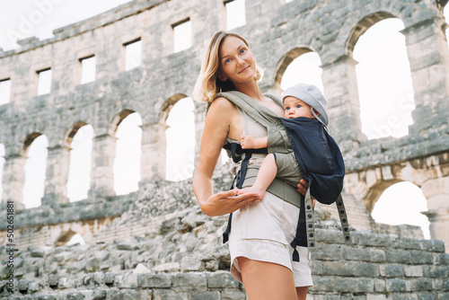 Tourist woman with child in Roman Amphitheater Arena like as Coliseum - famous travel destination in Pula, Croatia. Beautiful mother and baby in carrier on vacation in Europe. Family Active Travel.