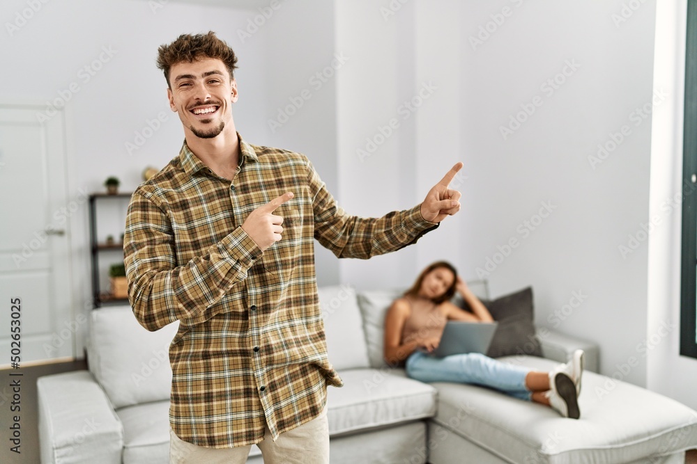 Young handsome man at the living room with girlfriend smiling and looking at the camera pointing with two hands and fingers to the side.