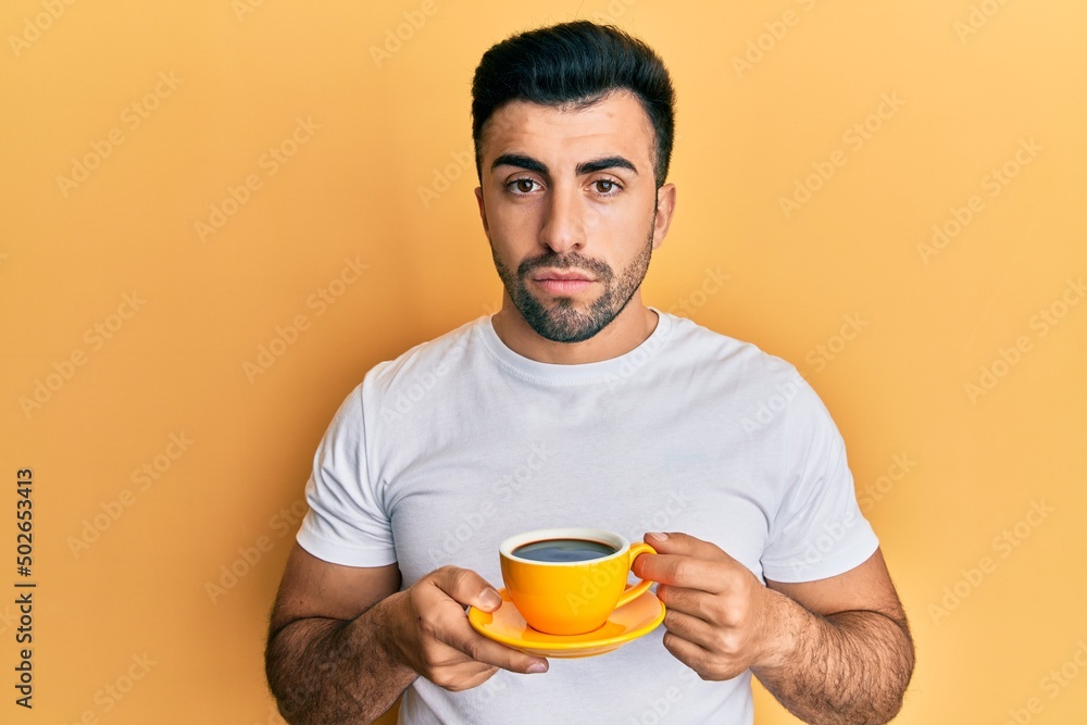 Young hispanic man drinking a cup of coffee relaxed with serious expression on face. simple and natural looking at the camera. Young hispanic man drinking a cup of coffee relaxed with serious expression on face. simple and natural looking at the camera.