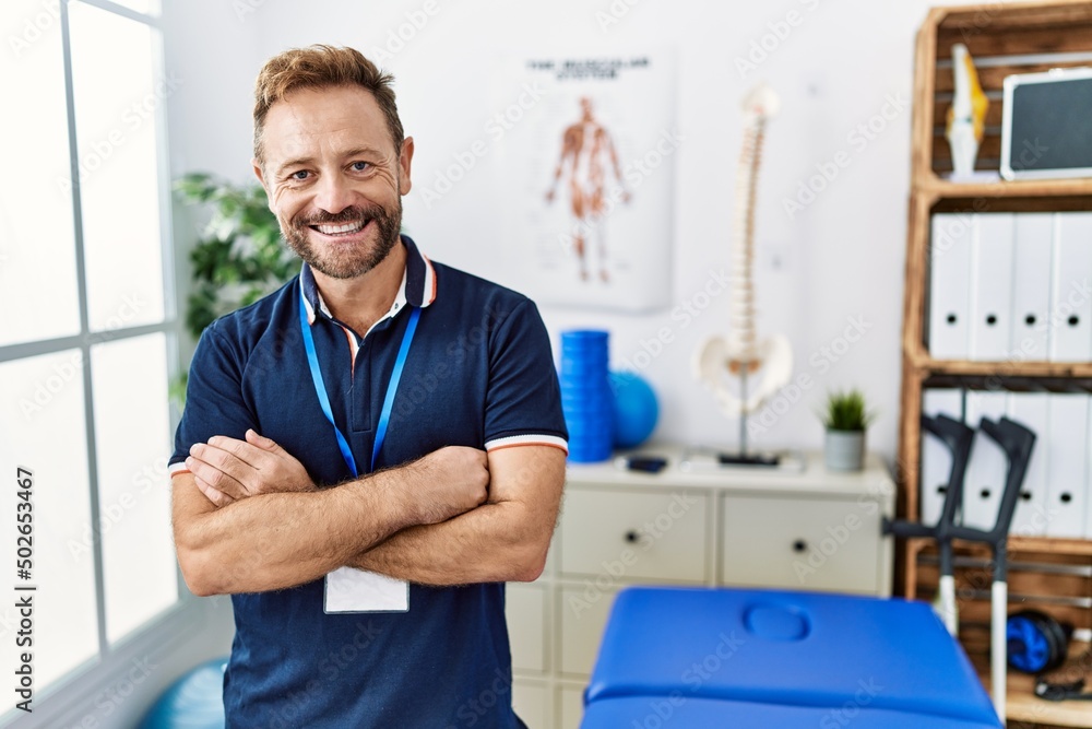 Middle age physiotherapist man working at pain recovery clinic happy face smiling with crossed arms looking at the camera. positive person. Middle age physiotherapist man working at pain recovery clinic happy face smiling with crossed arms looking at the camera. positive person.