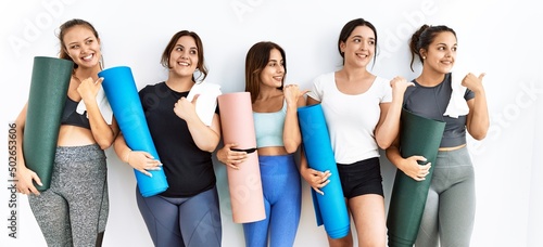Group of women holding yoga mat standing over isolated background pointing to the back behind with hand and thumbs up, smiling confident