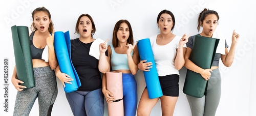 Group of women holding yoga mat standing over isolated background surprised pointing with finger to the side, open mouth amazed expression.
