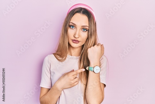 Young blonde girl wearing casual clothes in hurry pointing to watch time  impatience  looking at the camera with relaxed expression